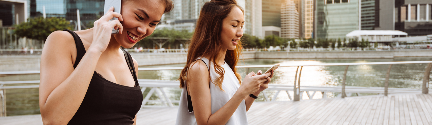 Two young women walk outdoors on a boardwalk by the water, surrounded by modern city buildings. One woman smiles while talking on her phone, and the other looks down, focused on her smartphone. It appears to be a sunny day.