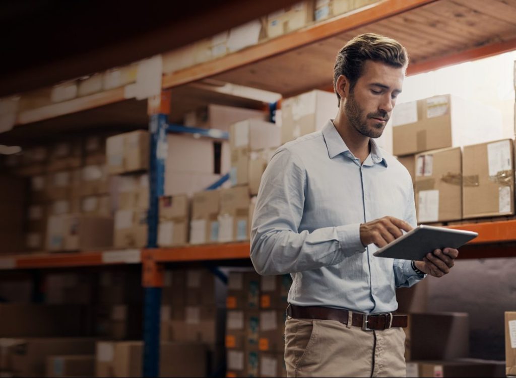 A man with short brown hair and a neatly trimmed beard stands in a warehouse, using a tablet. He wears a light blue dress shirt and beige pants. In the background are shelves filled with cardboard boxes and packages.