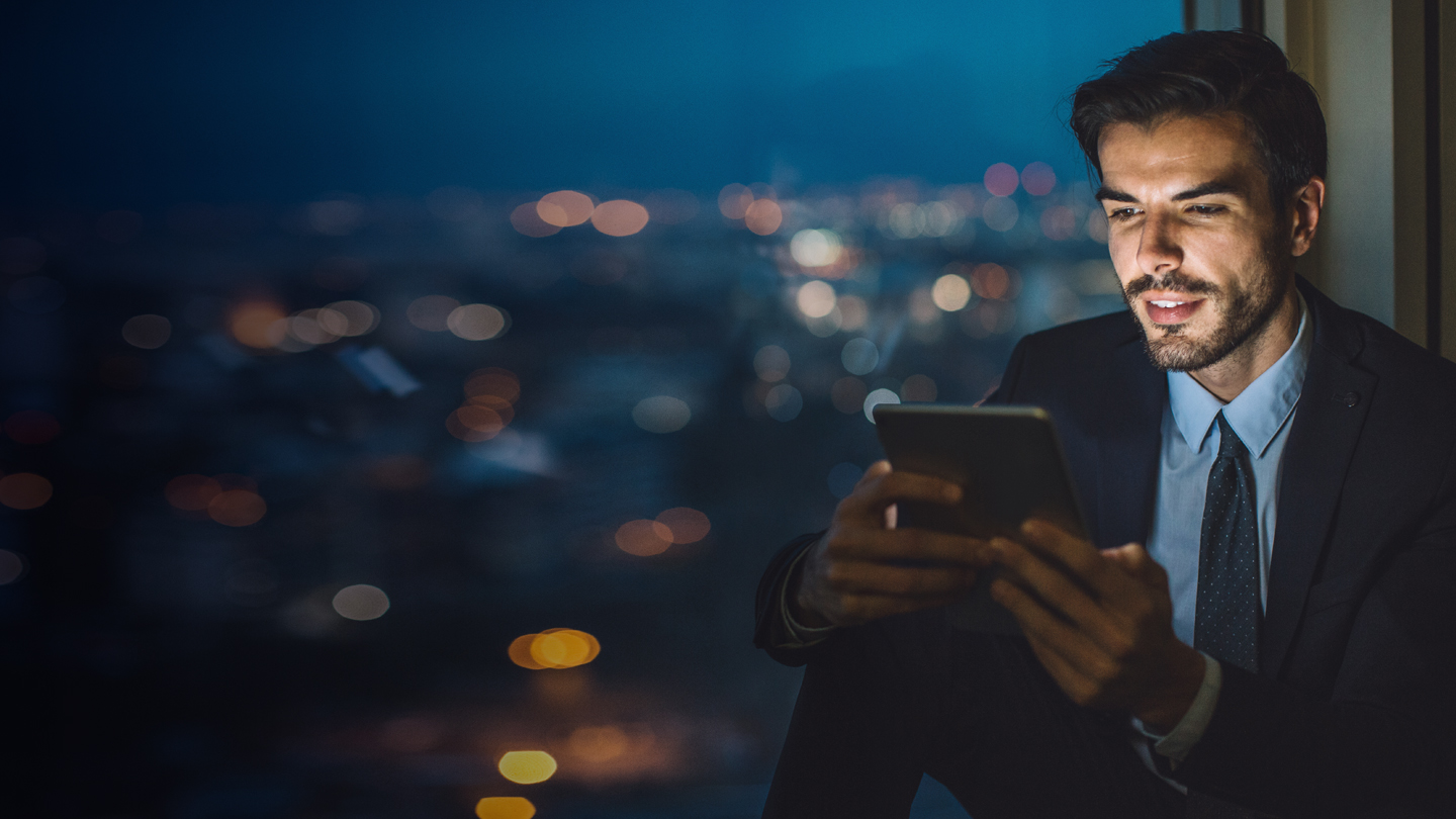 A man in a dark suit and tie sits by a window at night, holding a tablet and looking at the screen with a slight smile. The city skyline outside is blurred with colorful lights, creating a bokeh effect in the background.