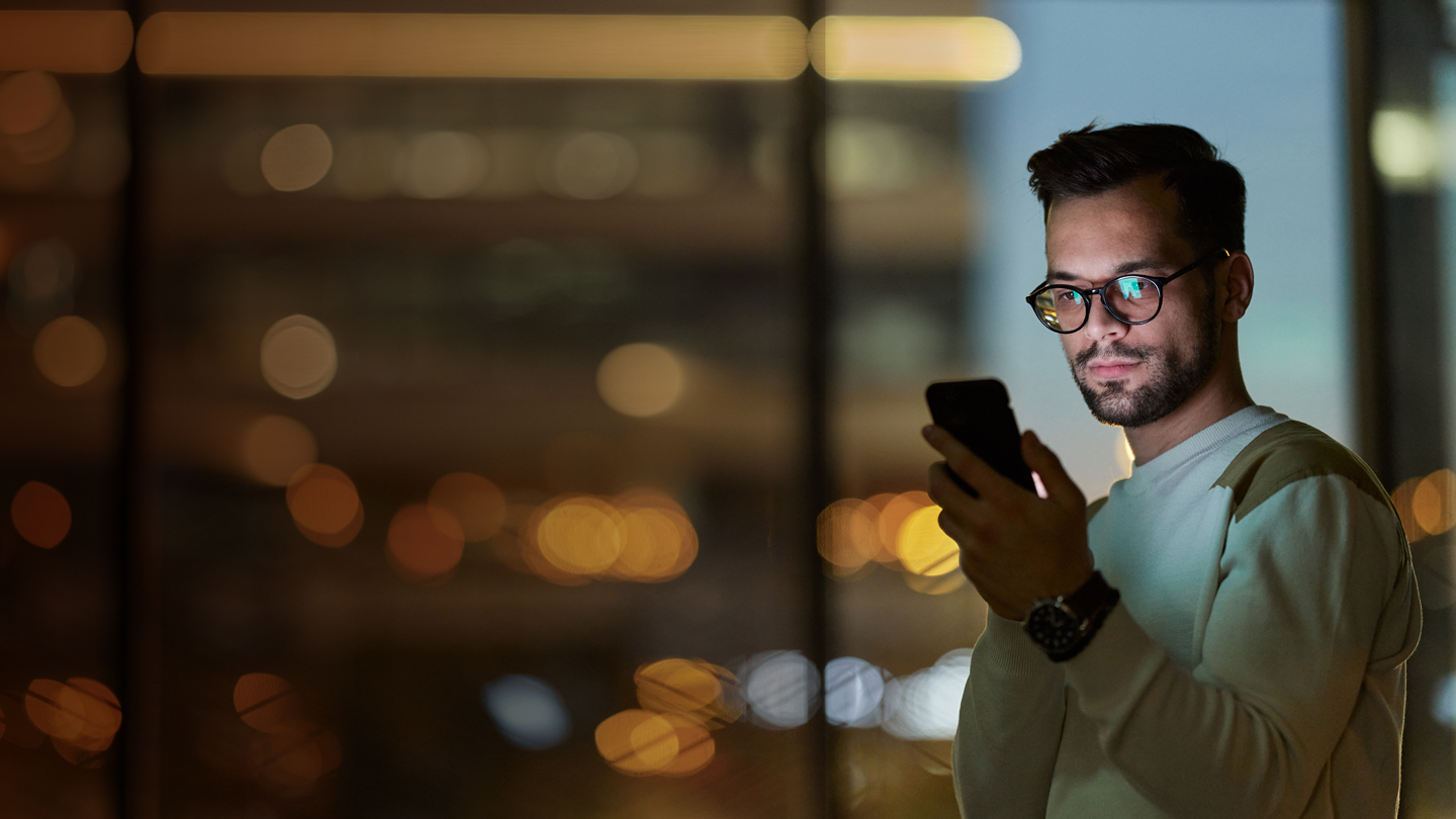 A man wearing glasses and a light sweater stands indoors at night, looking at his smartphone. Warm city lights and building windows are blurred in the background, creating a bokeh effect. The man appears focused on his device.