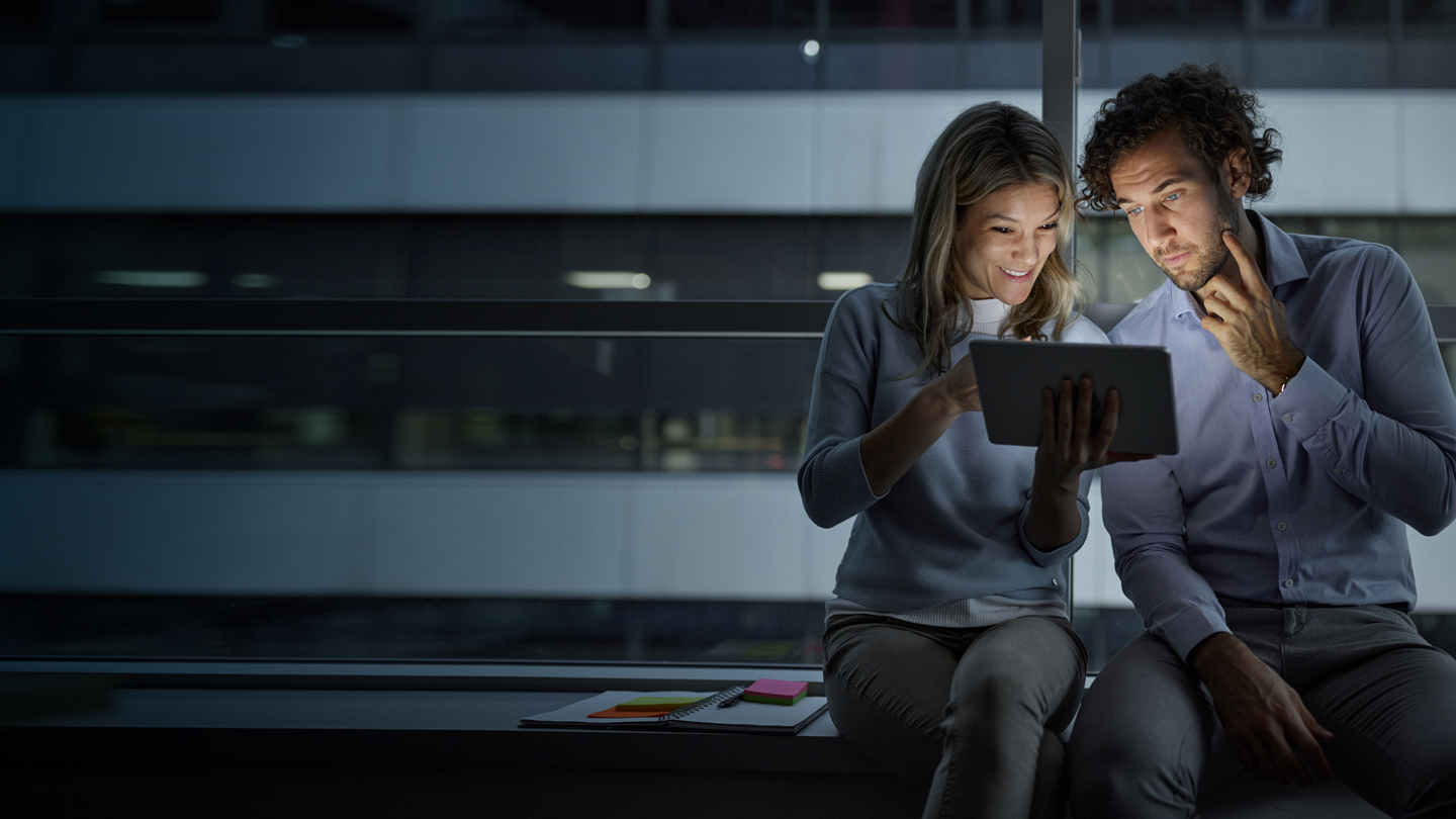 Two people sit side by side by a large window at night, illuminated by the glow of a tablet they are looking at together. The woman smiles and gestures at the screen, while the man looks thoughtful, resting his hand on his chin.