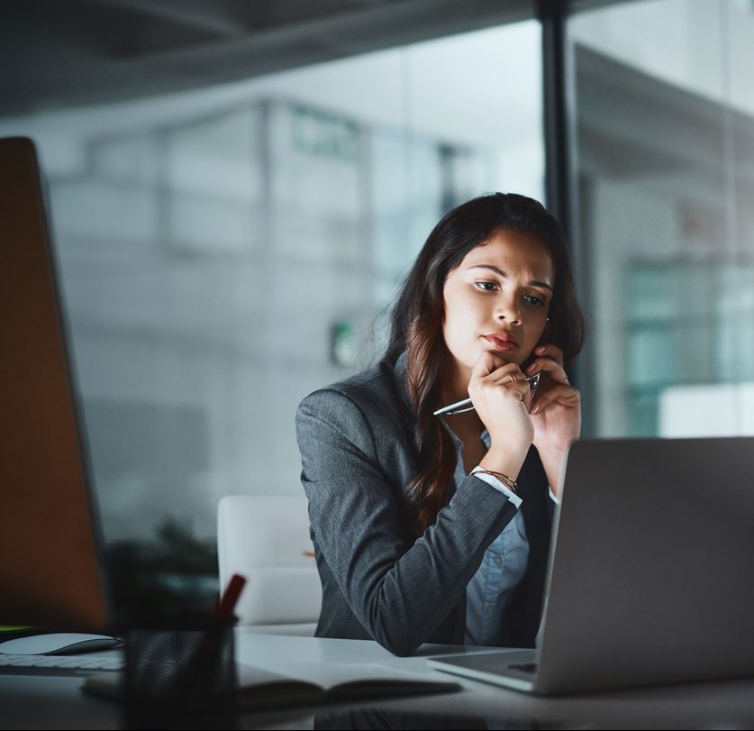 A woman in a gray blazer sits at a desk in an office, looking thoughtfully at her laptop screen. She holds a pen near her chin and rests her elbow on the desk. The background shows large glass windows and a modern office setting.