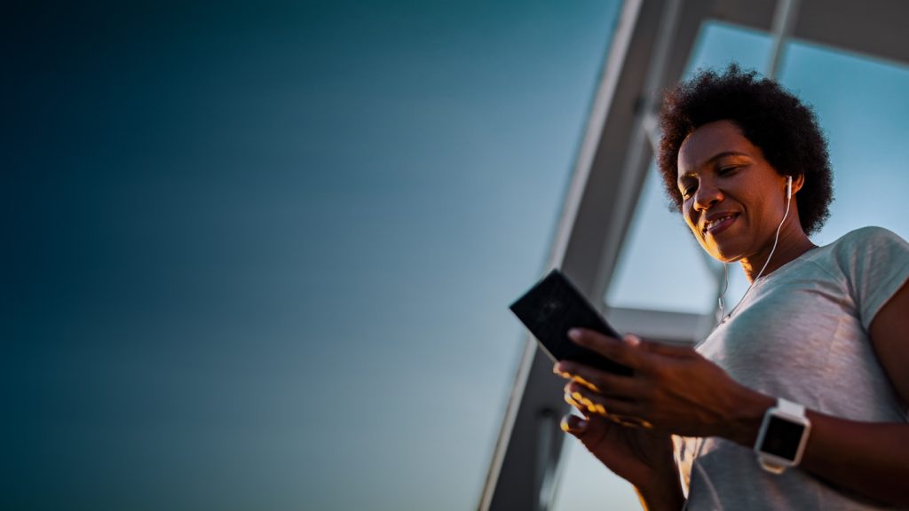A woman with short curly hair, wearing a white shirt and smartwatch, smiles while using her smartphone outdoors. She has earphones in, and the sky behind her is clear and blue, with part of a modern glass building visible.