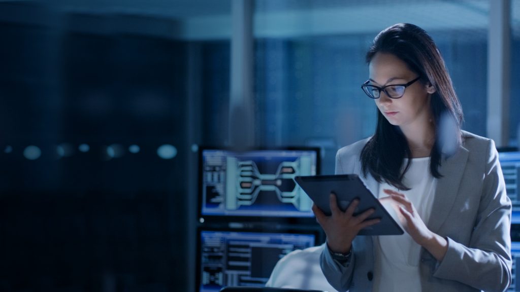 A woman in a gray blazer and glasses stands in a dimly lit office, looking at a tablet. Behind her are large monitors displaying data and graphics, suggesting a technology or cybersecurity environment. The scene has a modern, professional atmosphere.