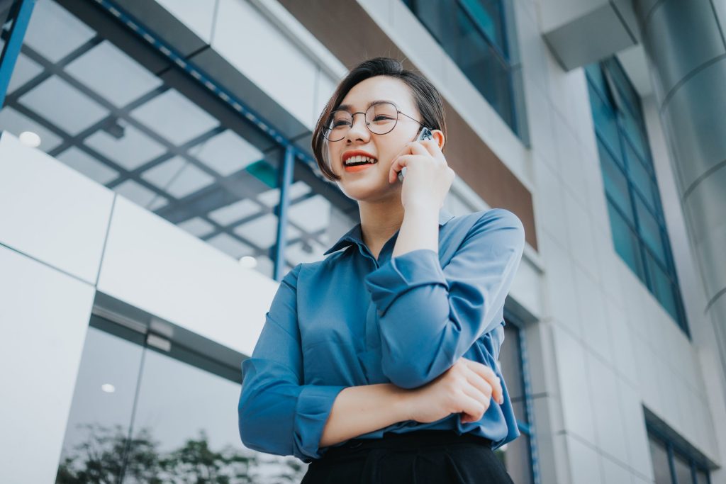A young person with short hair and glasses, wearing a blue shirt, stands outside a modern glass building, smiling and talking on a smartphone, with one arm crossed and a joyful expression. The background shows reflections and geometric architecture.