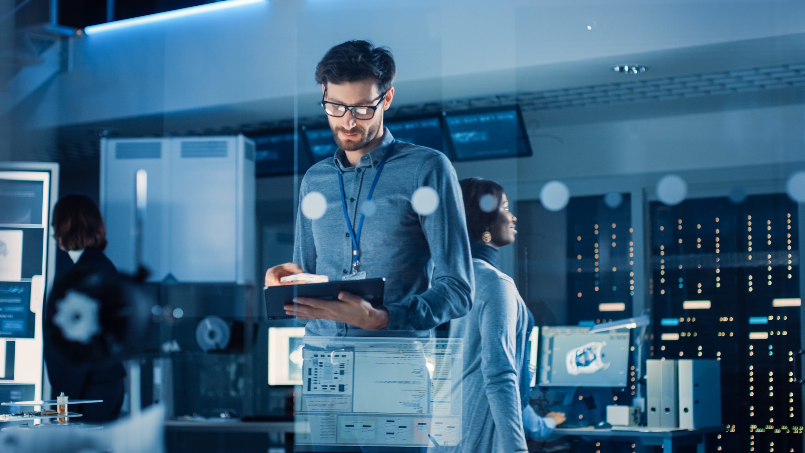 A man with glasses and a beard stands in a modern, high-tech office, looking at a tablet. Computer screens and server racks fill the background. A woman is seen working at a desk behind him. The scene has a blue, futuristic ambiance.
