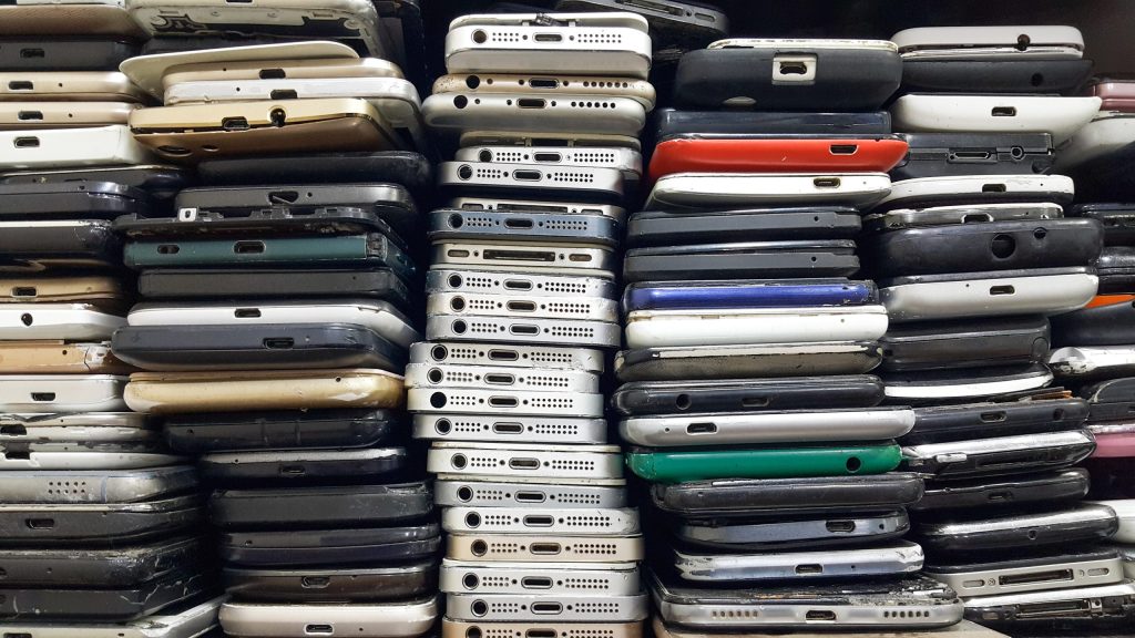 Stacks of old, used smartphones in various colors and conditions are piled closely together. Most devices are facing with their charging ports and speaker grilles outward, showing signs of wear and scratches.