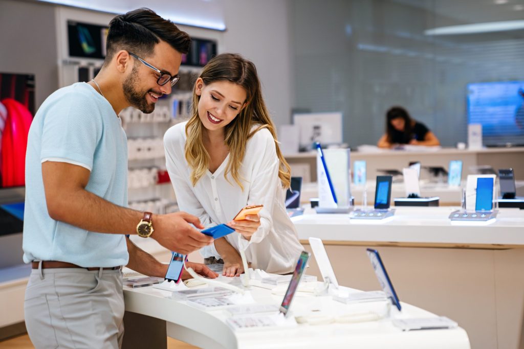 A smiling man and woman look at smartphones in a bright, modern electronics store. They seem interested and engaged, comparing two phones. Various smartphones are displayed on the counters around them, and another person is visible in the background.