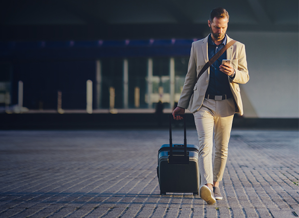 A man in a beige suit walks outside, pulling a rolling suitcase with one hand and looking at his phone in the other. He holds a coffee cup, has a trimmed beard, and sunlight casts shadows on the paved ground. The background is modern and blurred.