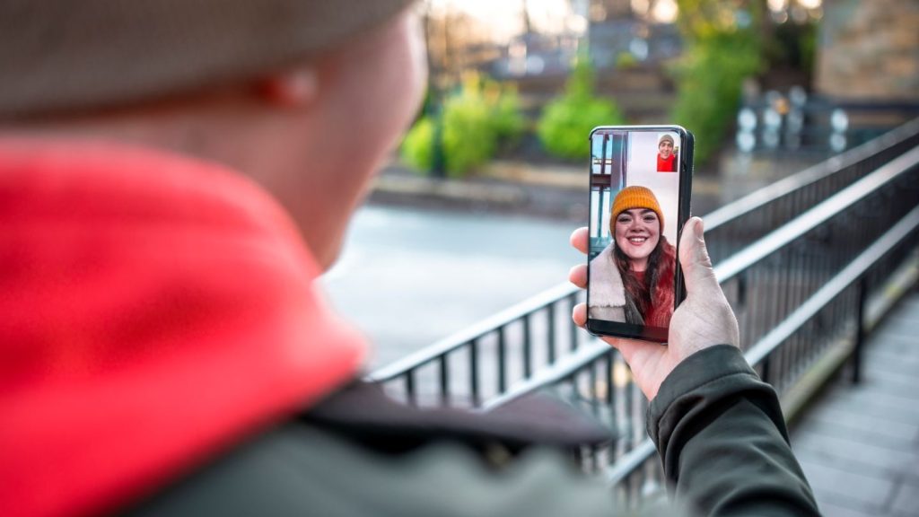 A person outdoors wearing a tan beanie and red jacket holds a smartphone, video calling someone who is smiling on the screen and also wearing a yellow beanie. The background shows a fence, pavement, and greenery in soft focus.
