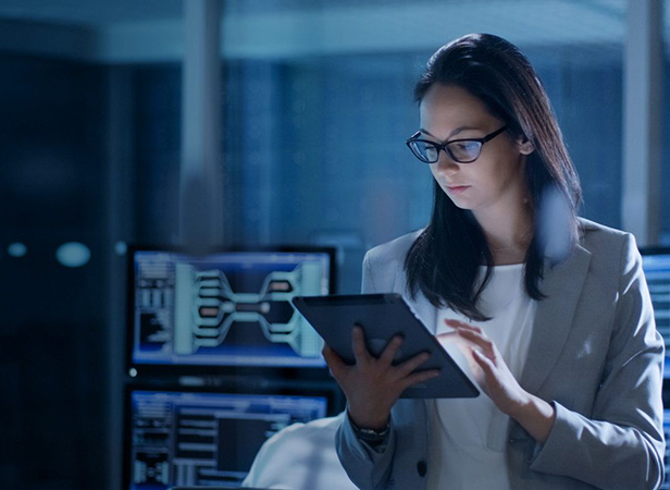 A woman in a gray blazer and glasses looks at a tablet in a dimly lit office. Behind her, computer screens display technical or scientific data, suggesting a technology or cybersecurity environment. The woman appears focused and professional.