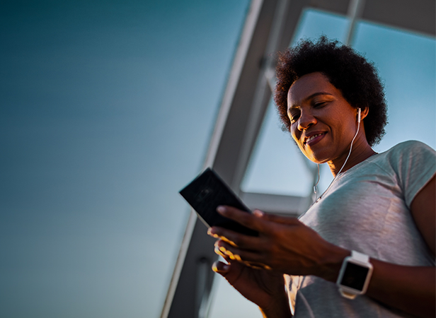 A person with short curly hair, wearing a white t-shirt, earphones, and a smart watch, smiles while looking at a smartphone outdoors. The sky is clear and blue, and modern glass architecture is visible in the background.