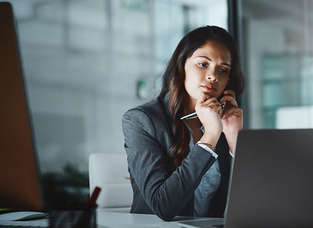 A woman in a business suit sits at a desk, holding a pen and talking on the phone while looking at her laptop screen. She appears focused and thoughtful, with a modern office environment visible in the background.