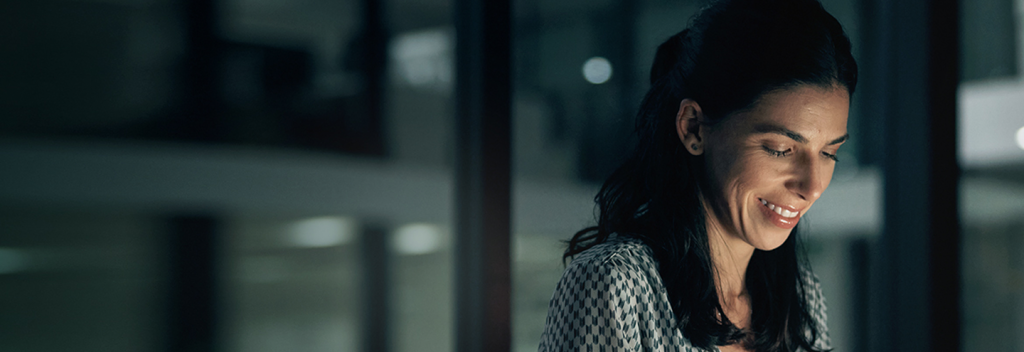 A woman with long dark hair smiles while looking down, illuminated by soft indoor lighting. She wears a patterned blouse and appears to be working or reading, with a blurred office background behind her.