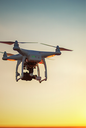 A white quadcopter drone with a camera hovers in the air at sunset, silhouetted against a colorful sky with warm orange and yellow tones near the horizon, fading into blue. The drone’s propellers are visible in motion.