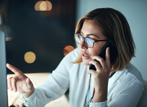 A woman with a bob haircut and glasses, wearing a white blouse, sits at a desk at night. She talks on the phone while looking at a computer screen, pointing at the monitor with her finger. The background is dimly lit with blurred lights.
