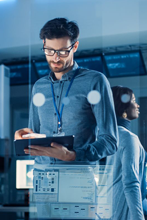 A man with glasses and a lanyard stands in a modern, high-tech office, looking at a tablet. Behind him, a woman faces away, both surrounded by computer screens, glass panels, and servers, suggesting a technology or cybersecurity environment.