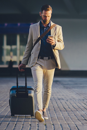 A man in a beige suit walks outside on a paved surface, pulling a rolling suitcase with one hand and looking at his phone in the other. He carries a shoulder bag and a coffee cup, with a modern building in the background. The lighting suggests early morning or evening.