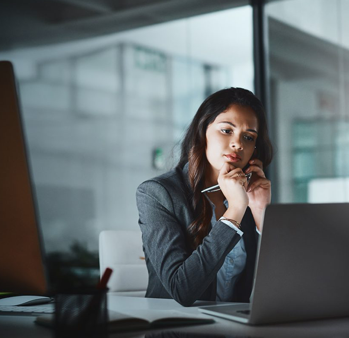 A woman in a business suit sits at a desk in a modern office, looking thoughtfully at her laptop screen. She holds a pen near her chin and rests her elbow on the desk. The background shows glass walls and a blurred office environment.