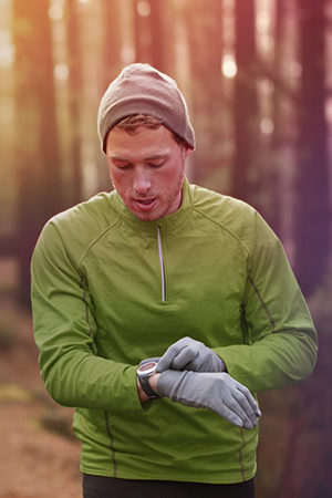 A man in a green long-sleeve shirt, gray gloves, and a gray beanie checks his smartwatch while standing on a forest trail, surrounded by tall trees and warm, golden sunlight filtering through the woods.