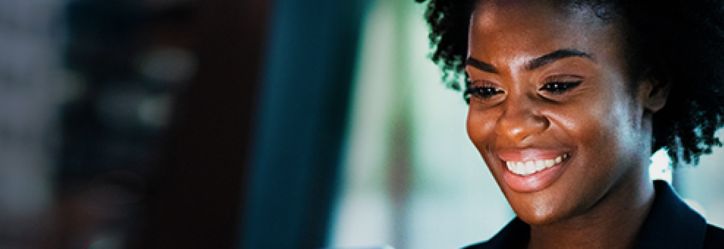A woman with curly hair smiles warmly while looking at a screen, possibly a computer or tablet. The background is softly blurred with cool tones, drawing attention to her cheerful expression and focused demeanor.