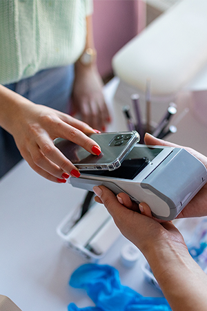 A person is paying with a smartphone using contactless payment at a card reader held by another person. Both are standing over a white table with salon tools and supplies visible in the background.