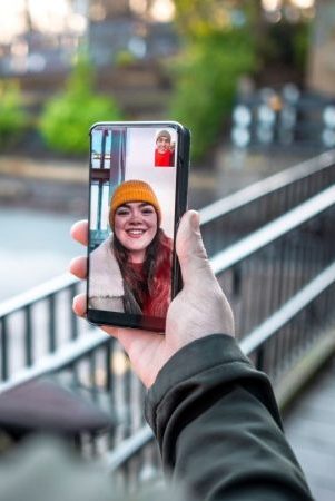 A person holds a smartphone while video calling someone outdoors. The phone screen shows a smiling woman in a yellow beanie and red scarf. The background includes a railing and blurred greenery, suggesting a park or urban setting.