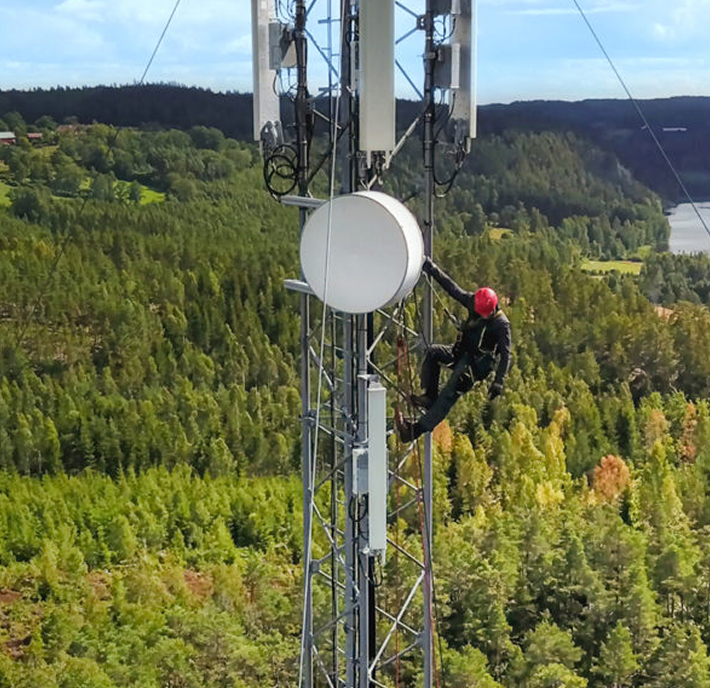 A worker wearing black clothing and a red helmet is climbing a tall metal telecommunications tower, surrounded by dense green forest and distant hills under a blue sky. The worker appears to be performing maintenance on the tower equipment.