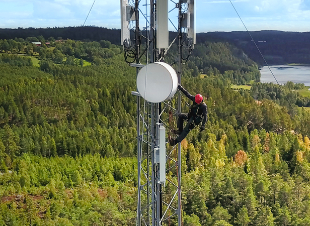 A worker wearing a red helmet and safety gear is climbing a tall metal cell tower in a green, forested area with a river or lake and hills in the background under a partly cloudy sky.