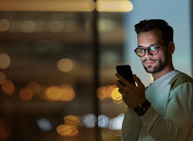 A man with glasses, a beard, and short dark hair is standing indoors at night, looking at his smartphone. He wears a light sweater and a wristwatch. Blurred city lights glow in the background through large windows.