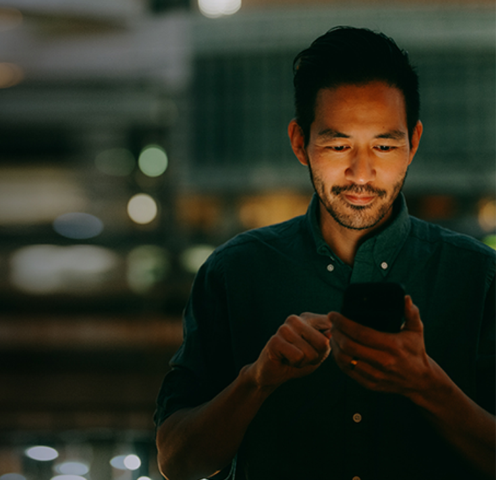 A man with short dark hair and trimmed beard stands indoors at night, softly illuminated by the light from his smartphone. He wears a dark button-up shirt and looks intently at his phone, with blurred city lights in the background.