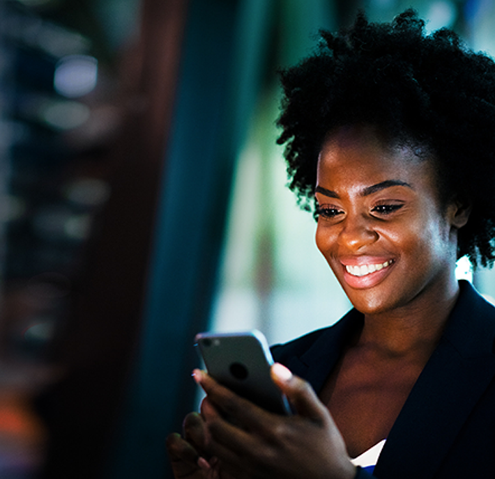 A woman with curly hair and a dark blazer smiles while using a smartphone. She is indoors, illuminated by soft, focused lighting, with a blurred, modern background suggesting an office or workspace environment.