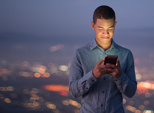 A young man in a light blue shirt stands outdoors at dusk, smiling as he looks at his smartphone. The city lights behind him are blurred, creating a colorful bokeh effect against the darkening sky. His face is softly illuminated by his phone’s screen.