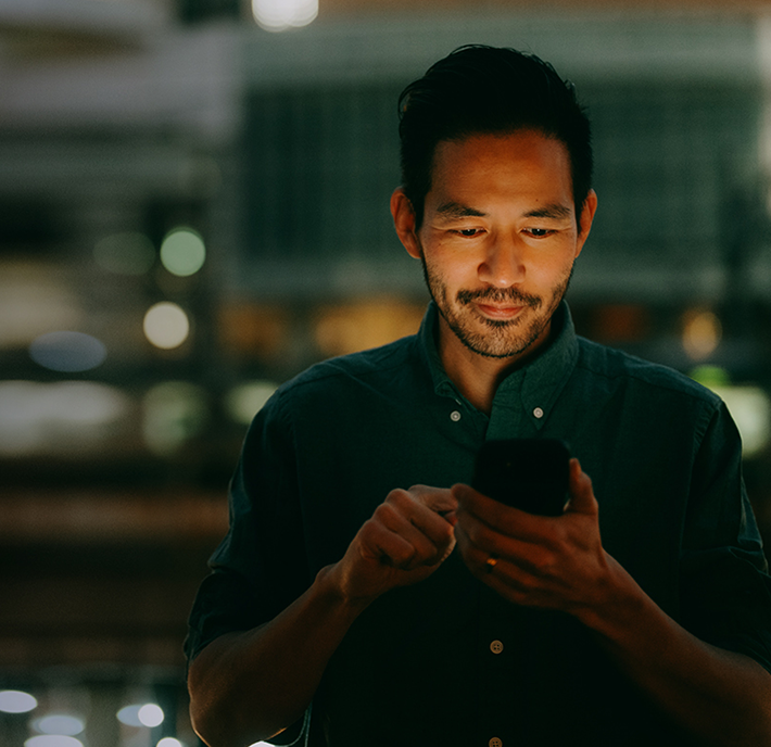 A man with dark hair and a trimmed beard stands indoors at night, illuminated by his smartphone. He wears a dark shirt and looks at the phone with a slight smile. The blurred city lights in the background create a warm, bokeh effect.