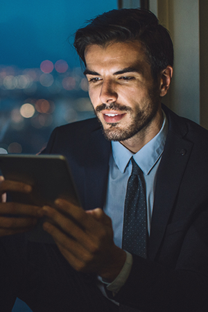A man in a suit sits by a window at night, smiling as he looks at a tablet. The city lights outside are blurred, creating a bokeh effect. The scene suggests he is working or relaxing in a modern office or apartment.
