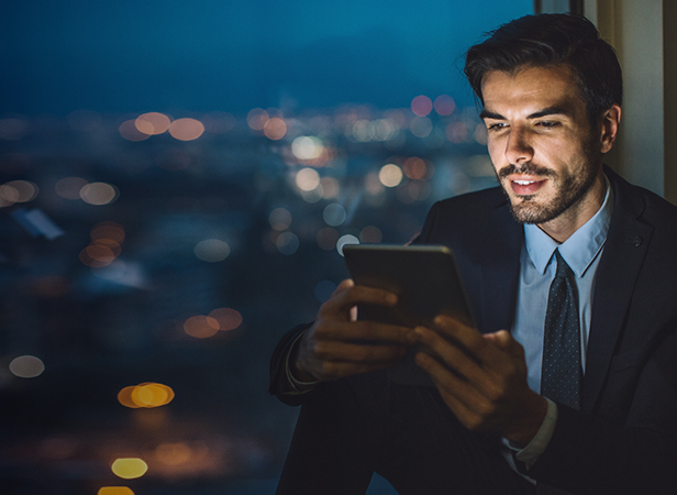 A man in a suit sits by a window at night, holding and looking at a tablet. City lights create a bokeh effect in the background, giving a modern, urban atmosphere. The man appears engaged and focused on the device’s screen.