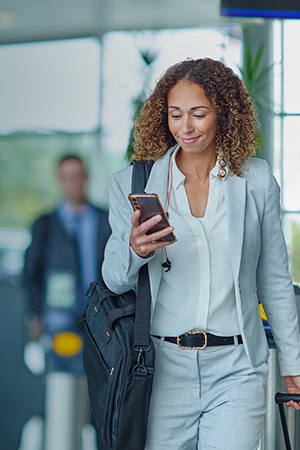 A woman in a light gray suit, carrying a black shoulder bag, smiles while looking at her phone in a bright, modern building. Two blurred people are in the background near entry gates. Sunlight streams through large windows.