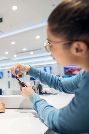 A man with a beard and glasses smiles while leaning on a white counter in a tech store. A woman in a blue top demonstrates a smartphone to him. The background is brightly lit, with shelves and screens visible, suggesting an electronics retail setting.