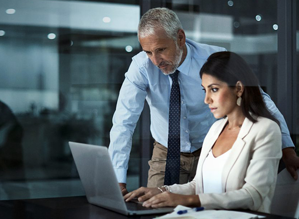 A woman in a white blazer sits at a desk, focused on her laptop. A man with gray hair and a beard, wearing a blue shirt and tie, stands beside her, looking at the screen. They appear to be in a modern office setting at night.