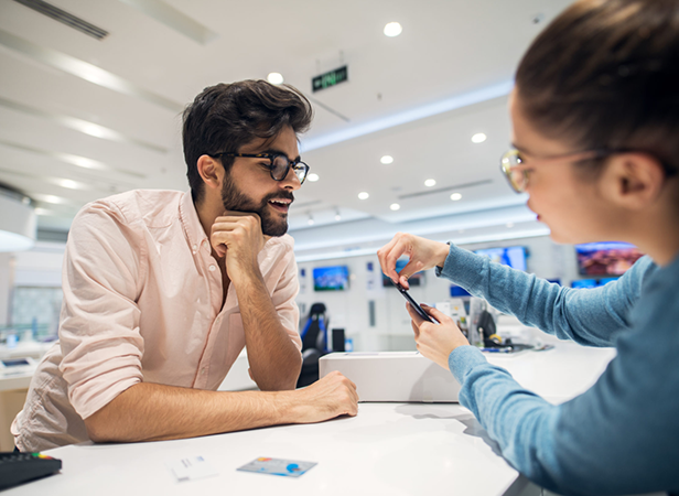 A man with a beard and glasses leans on a counter, smiling as a woman in a blue sweater shows him a smartphone. The setting appears to be a modern electronics store with bright lighting and shelves in the background. Credit cards are on the counter.