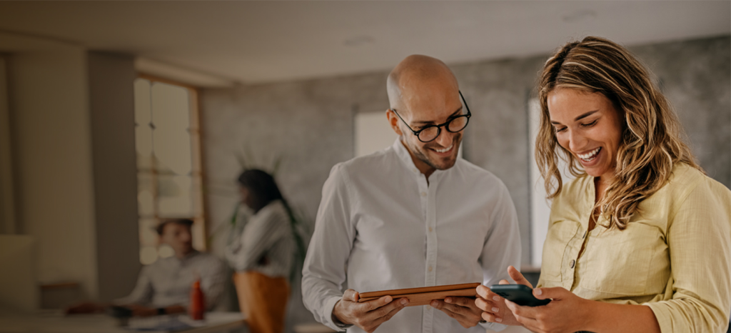 Two colleagues stand indoors, smiling and looking at a smartphone together—perhaps reviewing GSMA Services. The man, bald and wearing glasses, holds a tablet. In the blurred background, two more people converse near a window in a bright, friendly scene.