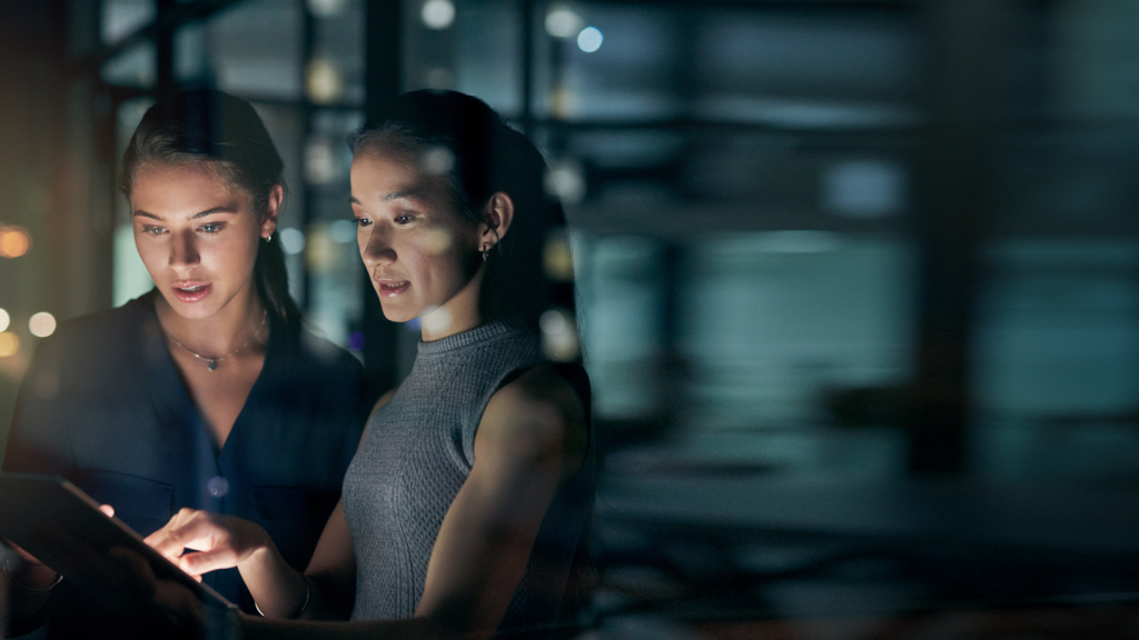 Two women stand together in a dimly lit office, intently examining a glowing tablet. One points at the screen, highlighting TAC Allocation insights from GSMA Services, while the other observes. Blurred office lights frame their focused discussion.