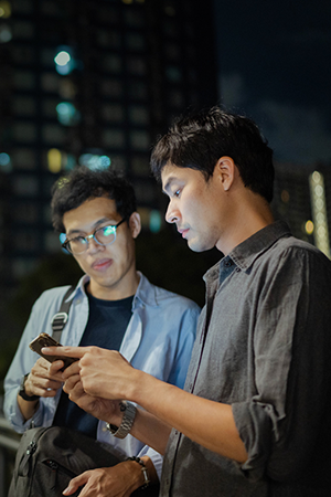 Two men stand outdoors at night in an urban setting. One man wears glasses and looks at his phone, while the other, lit by the phone’s screen, holds a bag and watches. Tall buildings with lit windows are visible in the blurred background.