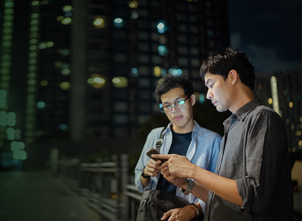 Two young men stand outside at night, illuminated by city lights and the glow of a smartphone. One man in glasses looks at the phone while the other holds it. Tall buildings with many windows are blurred in the background.