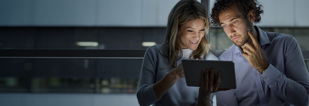 A woman and a man stand close together indoors, looking intently at a digital tablet the woman is holding. The man touches his chin thoughtfully. Both appear engaged and focused. Large windows and a blurred office building are in the background.
