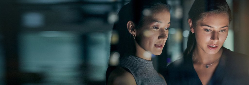 Two women stand closely together in a dimly-lit setting, looking intently at a glowing screen or monitor. Reflections and blurred lights are visible on the glass in front of them, creating a focused and contemplative atmosphere.