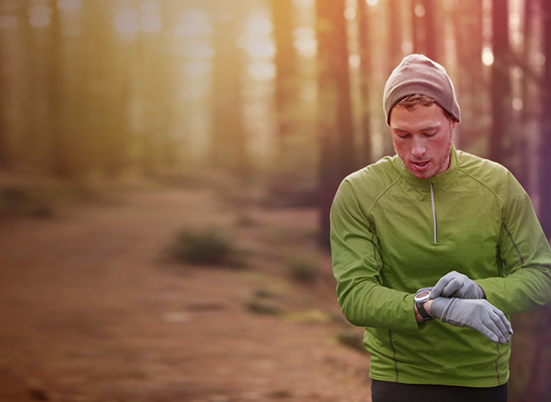 A man wearing a green jacket, gray gloves, and a beanie stands in a sunlit forest, looking down at his smartwatch. The forest path behind him is blurred, and soft sunlight filters through the tall trees, creating a warm, serene atmosphere.