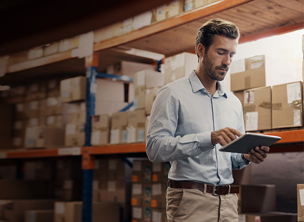A man in a light blue shirt uses a tablet in a warehouse filled with shelves and brown cardboard boxes. He stands in front of stacked packages, appearing focused as he checks or inputs information on the device.