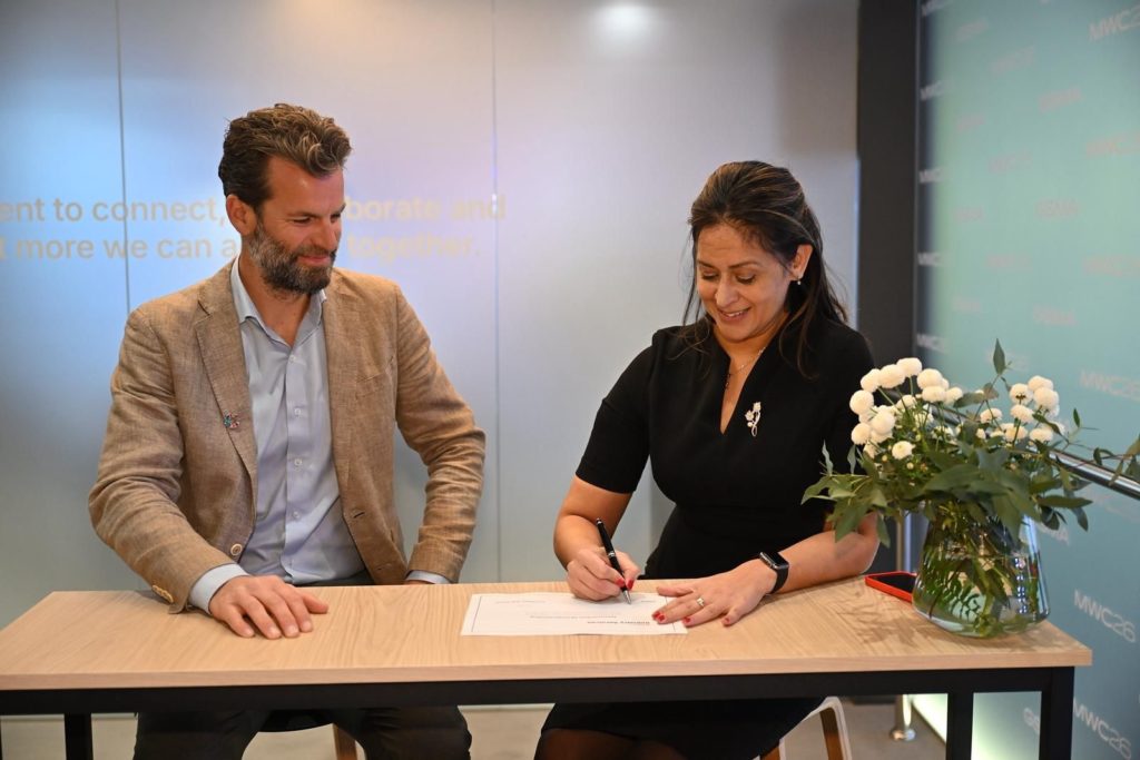 A man in a beige blazer and blue shirt sits beside a woman in a black dress who is smiling and signing a document at a light wooden table. A vase of white flowers and a red smartphone are on the table. The background is softly lit with blue-gray tones.