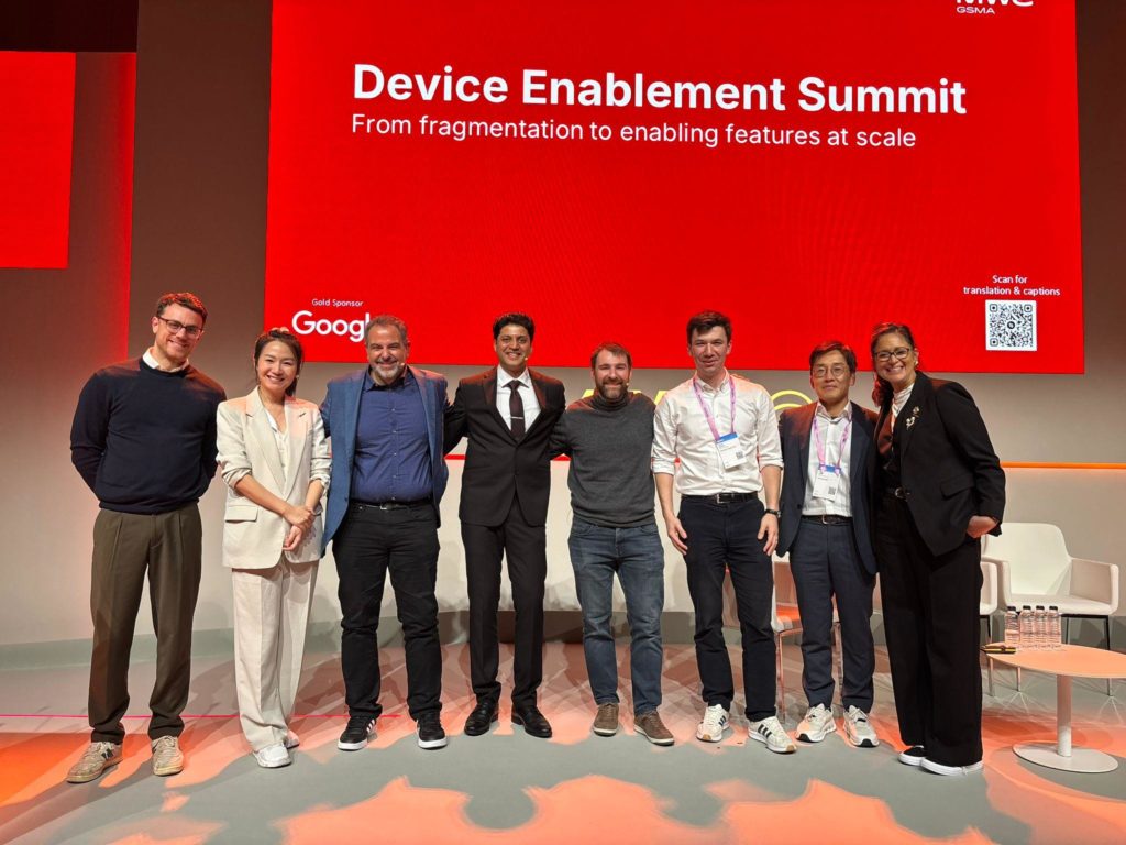 Eight people stand side by side on stage, smiling at the camera. Behind them is a large red screen displaying “Device Enablement Summit: From fragmentation to enabling features at scale,” with logos for Google and a QR code on the right.